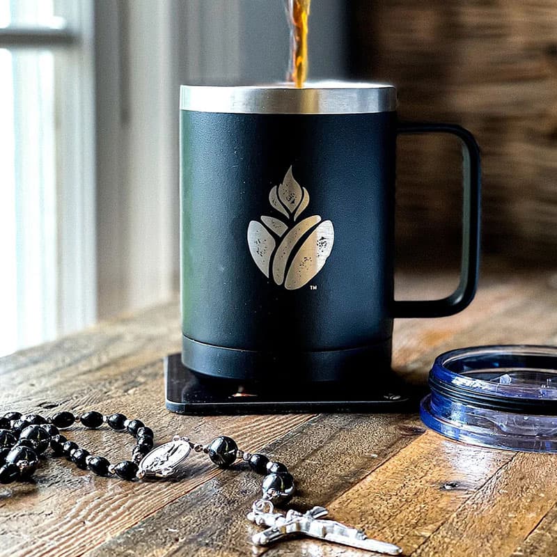 Black Catholic Coffee travel mug on a wooden surface, with coffee being poured into it.