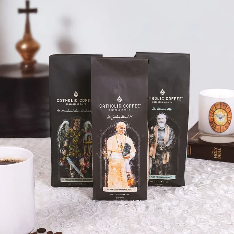 Three bags of Catholic Coffee with religious figures on the packaging, next to a white mug and a bible.